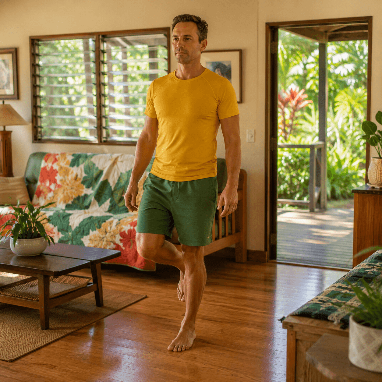 A local Caucasian man in a mustard-yellow t-shirt stands on one leg in his living room, focused and calm, arms relaxed.